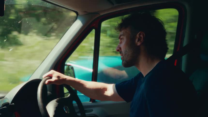 The video shows a man with a beard and sunglasses driving a red camper van he looks out the side window as he drives and at one point he takes his sunglasses off
