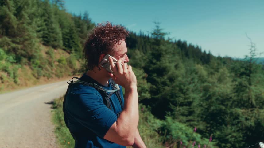 A man on a mountain bike is using his phone he is outdoors in a hilly forested area with a clear sky