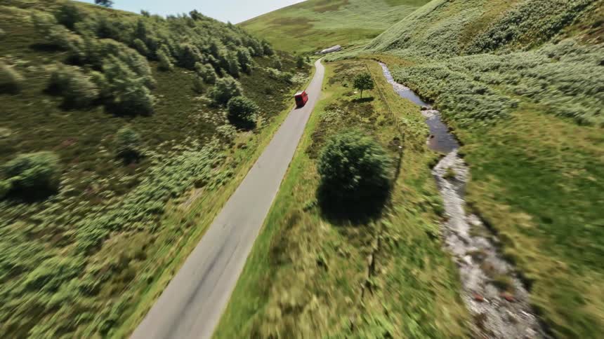 A red vehicle travels along a winding rural road bordered by green hills and a narrow stream