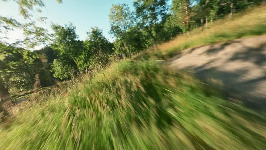 A solo mountain biker rides along a scenic sunlit dirt trail through a lush green countryside with scattered trees and a clear blue sky