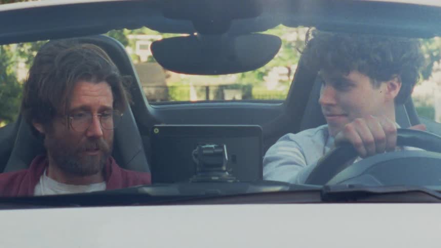 Two men are sitting in a convertible car and are nodding their heads while listening to music