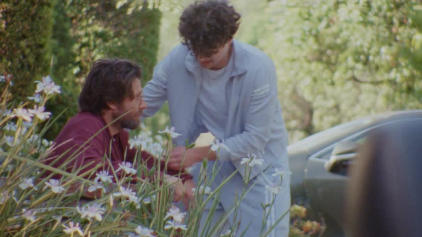 Young man helping a bearded man with glasses stand up among white flowers
