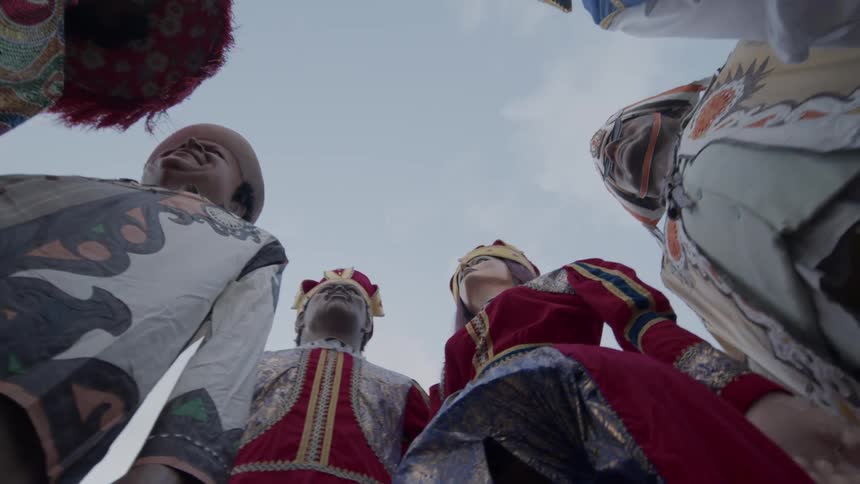 A group of people in colorful elaborate costumes and masks stand in a circle viewed from a low angle against the sky