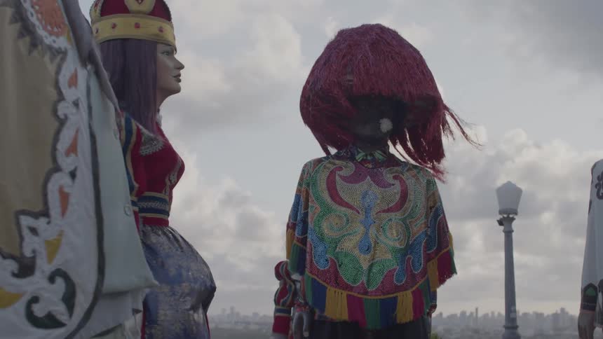 Giant puppets dressed in colorful clothes appear under a cloudy sky