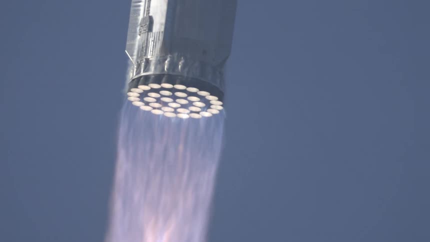The underside of a rocket in flight displays dozens of bright engine nozzles firing releasing plumes of hot exhaust against a clear blue sky
