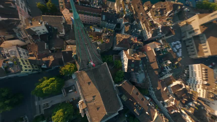 An aerial view captures a historical clock tower with a steeple surrounded by a city and a river with the sun setting behind the hills in the distance