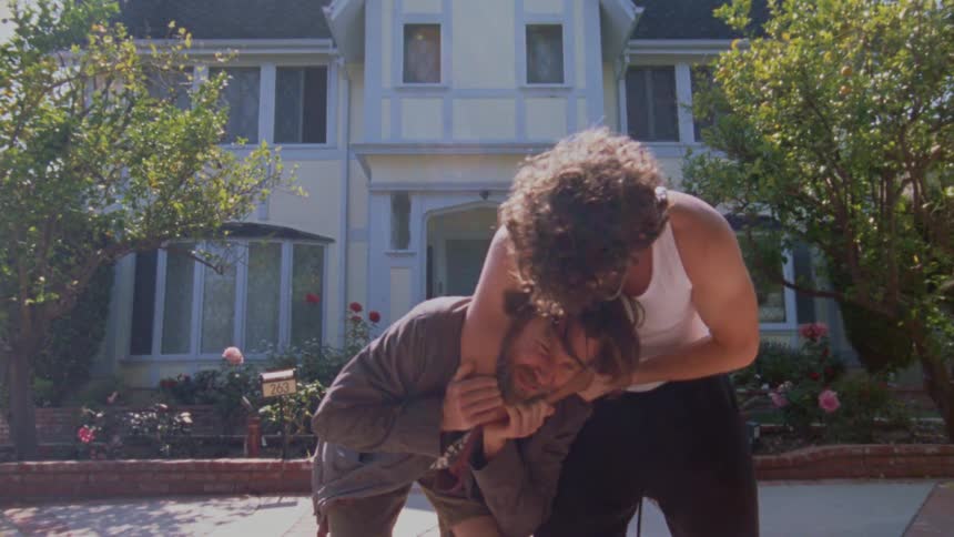 Two young men are wrestling and playing on a brick driveway in front of a house one man wearing a white tank top has the other in a headlock