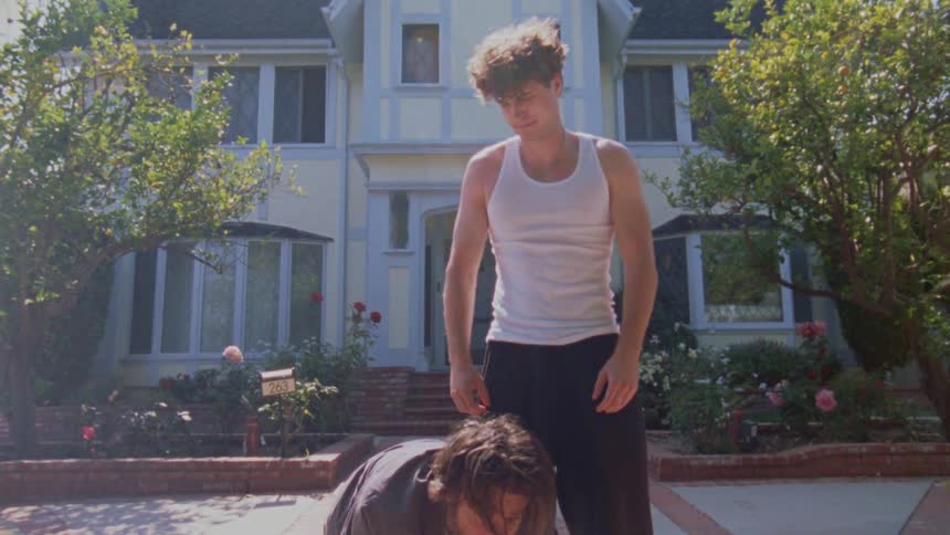 Two young men are wrestling and playing on a brick driveway in front of a house one man wearing a white tank top has the other in a headlock