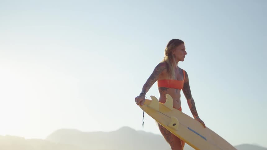 A blonde woman with tattoos is holding a yellow surfboard she is standing on a beach with a mountain range in the background
