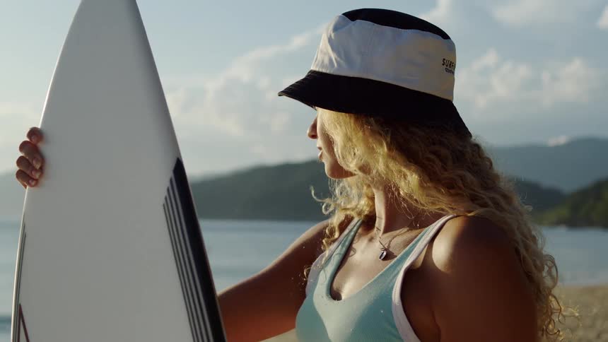 Surfer wearing a hat holds board on the beach looking at the sea