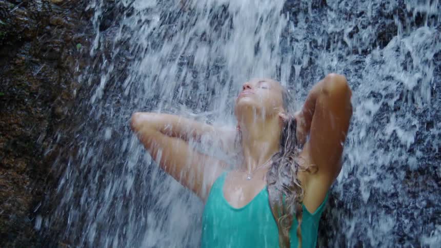 A woman in a turquoise swimsuit stands under a waterfall with her hands in her hair enjoying the cascade