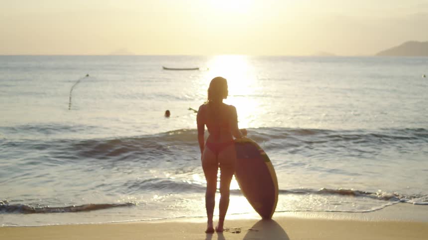 A female surfer stands on the beach watching the sun set