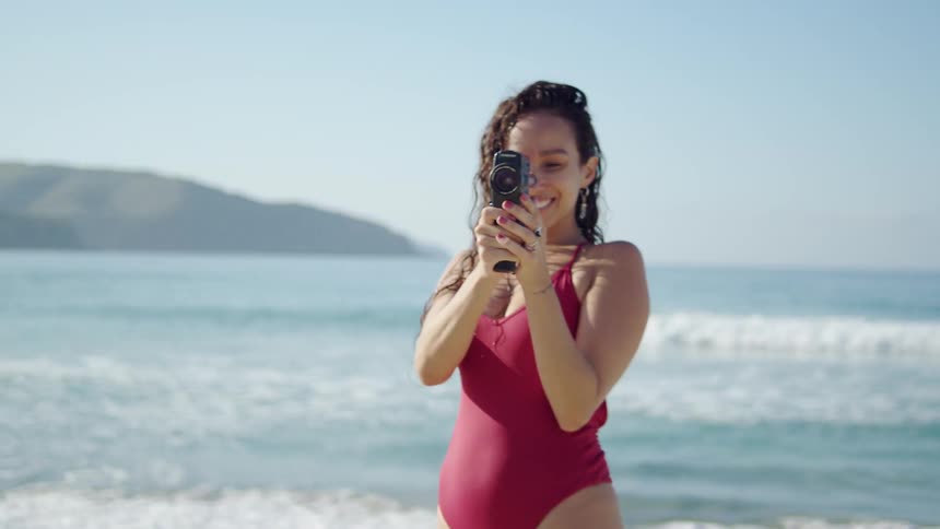 A young woman wearing a red swimsuit has fun with a camera on the beach at midday