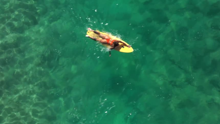 An aerial view looks down on a person wearing a red swimsuit lying on a yellow surfboard in clear shallow water the water is a brilliant green with sunlight dancing across its surface