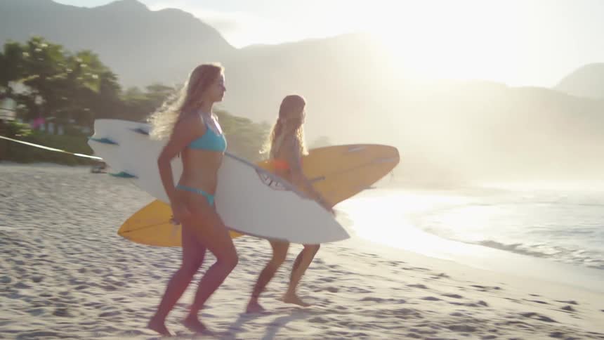 Two women walk on the sand carrying surfboards toward the sea