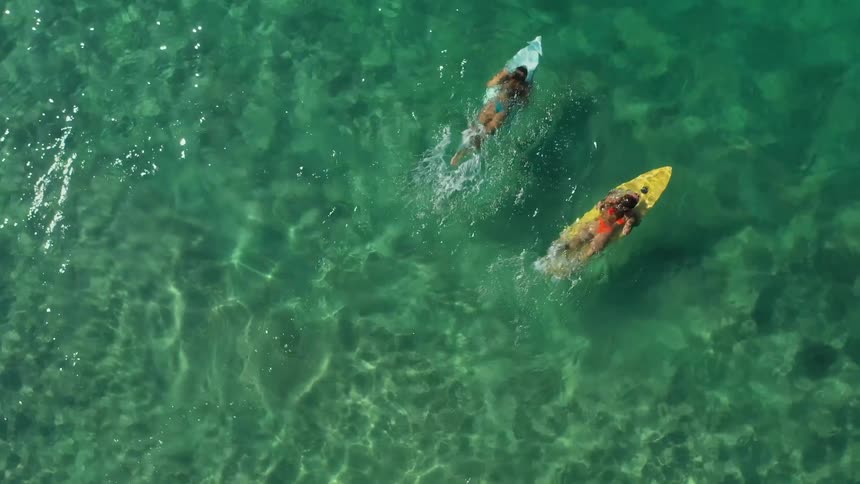 An aerial view shows two people on surfboards floating on the clear turquoise water they are lying on their boards one on a white board and the other on a yellow one