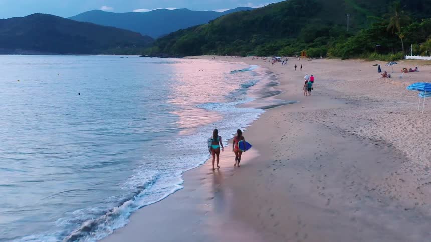 Two women walk along a sandy beach at sunset one carrying a surfboard with gentle waves and mountains in the background