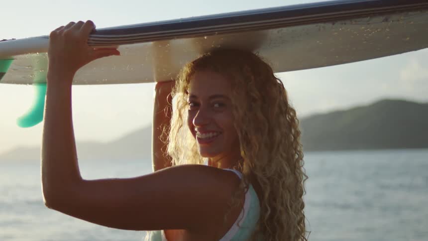 A medium shot from the side shows a person with long curly blonde hair carrying a surfboard on their head the sun is low and bright creating a lens flare and backlighting the person against the ocean and a distant shoreline