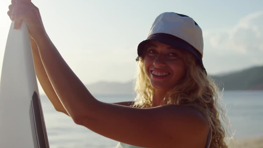 Teenage girl stands on the beach holding her surfboard as she turns and smiles