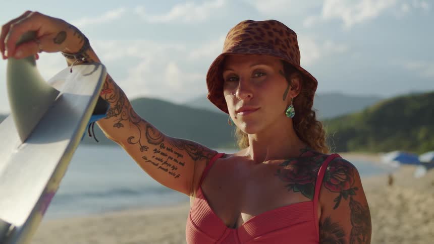 A woman with a leopard print bucket hat and a pink bikini top is on the beach she has multiple tattoos on her arms and shoulder and is holding a surfboard