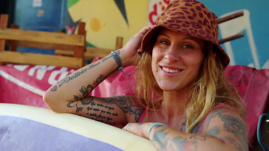 Smiling woman wearing a patterned hat leans on a surfboard