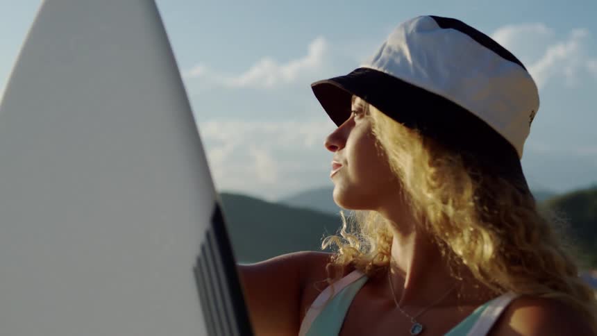 Woman with a black and white hat next to a surfboard on the beach