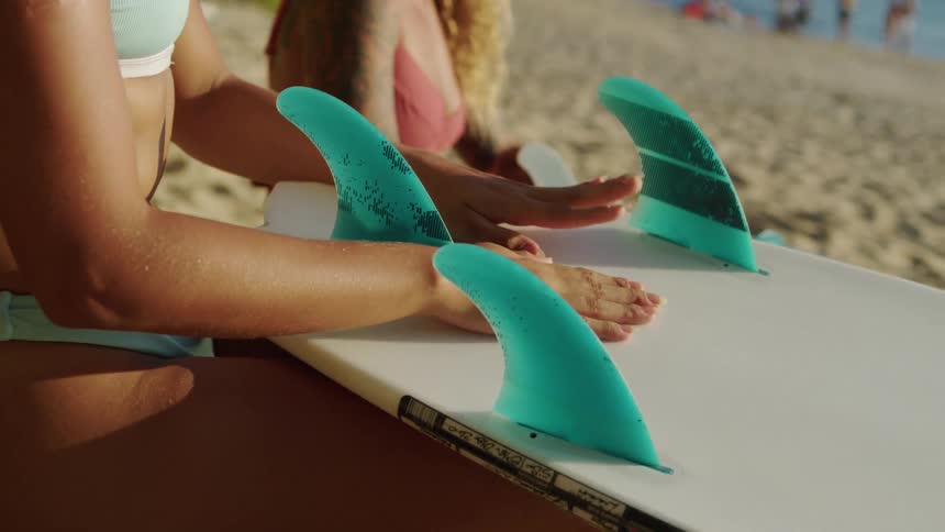 Two young women in bikinis sitting on the beach with surfboards in their laps beating a rhythm with their hands on the surfboards