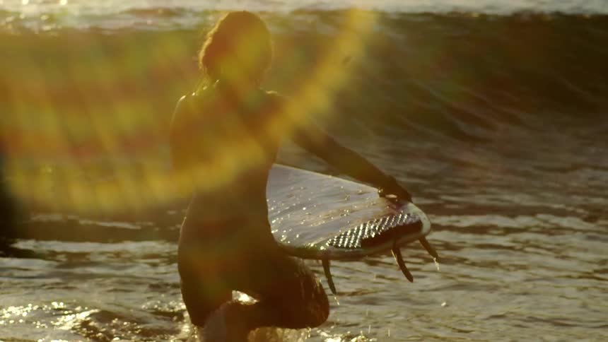 The silhouette of a woman holding a surfboard and walking into the sea with a wave breaking next to her