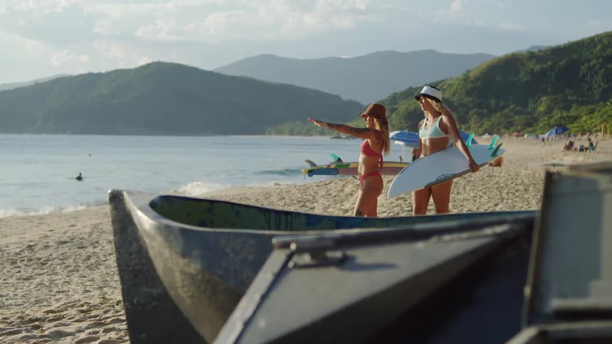 The video shows a person in a pink bikini and a person in a white bikini with a hat on standing on a sandy beach both are holding surfboards and looking out at the water and the mountains in the distance