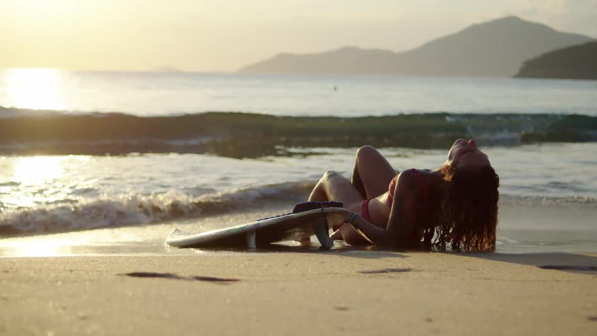 Woman lying on the sand at sunset with a surfboard beside her