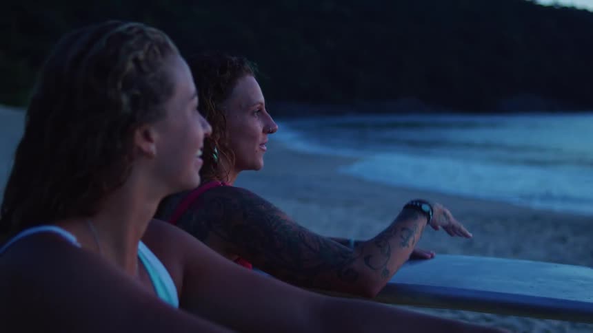 Two women talk on the beach while holding surfboards