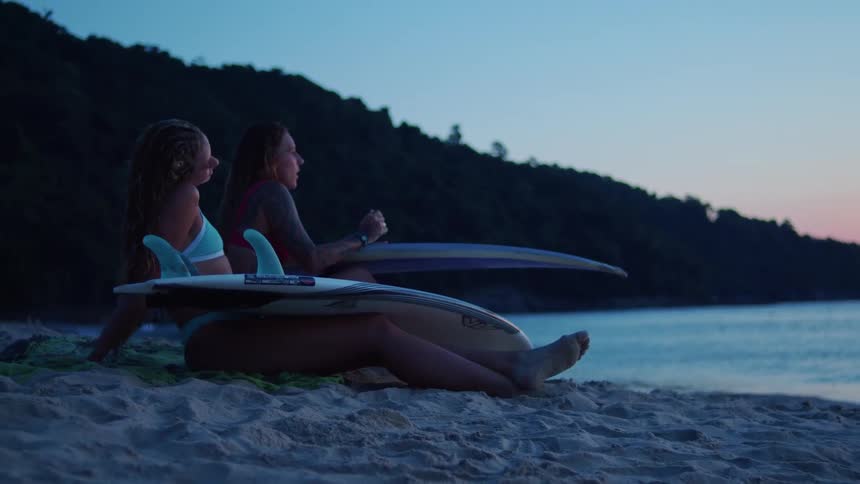 Two friends sit on the beach with surfboards at sunset