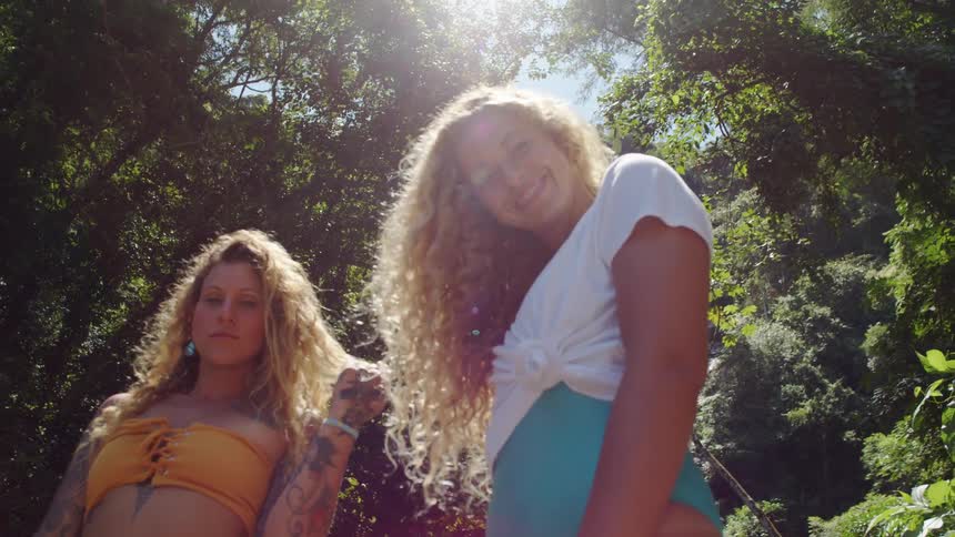 In the video two women are posing outdoors in a lush green environment the one on the right with long blonde curly hair is smiling and wearing a white tshirt tied at the front and what appears to be a blue or turquoise skirt or shorts the woman on