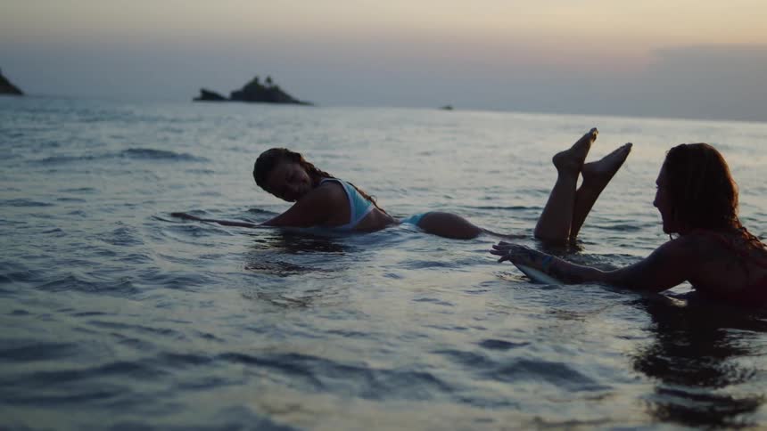 Two women lying on surfboards in calm ocean water on a day with the sun shining in the background