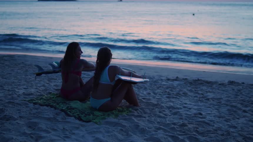 Two women sit on the beach discussing while each holds a surfboard at sunset