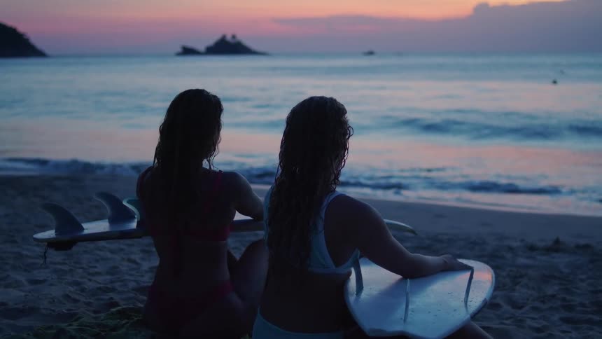 Silhouette of surfer girls sitting on sand near shoreline looking out to the ocean