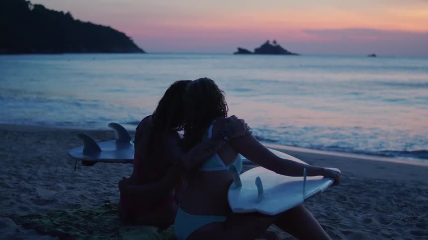 The video shows two young women sitting on a sandy beach at dusk with their backs to the camera they are both holding surfboards and are silhouetted against a beautiful sunset sky with a mix of purple pink and orange hues in the distance the ocean a