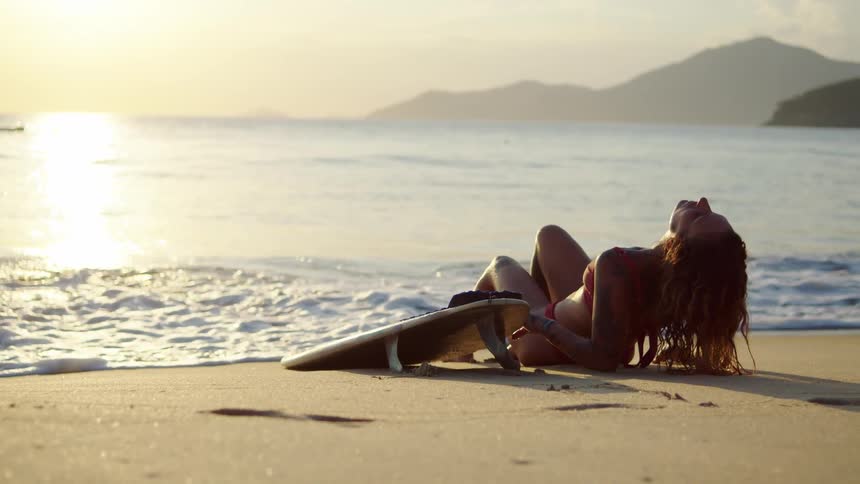 The video shows a woman in a red bikini lying on a sandy beach next to a surfboard at sunset she is positioned on her stomach looking out at the ocean while the sun sets over the water and a mountain range in the distance