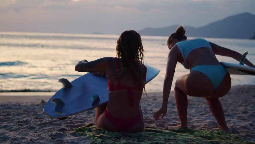 The video shows a woman in a pink bikini and another in a blue bikini sitting on a sandy beach both women are holding surfboards with the woman in pink holding hers in a way that blocks her torso from view they are looking out at the ocean as the sun s