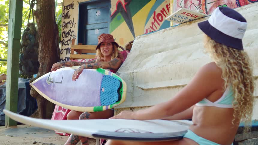 Two women are sitting outdoors with surfboards and laughing one woman is wearing a bucket hat with a leopard print and the other is wearing a white and blue bucket hat
