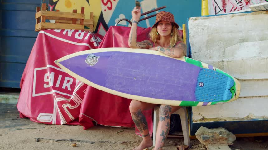 The video shows a medium shot of a person with tattoos and a bucket hat sitting on a stool and holding a purple surfboard with green edges the background is a vibrant cluttered scene with red and yellow fabrics and graffitilike art on the walls the w
