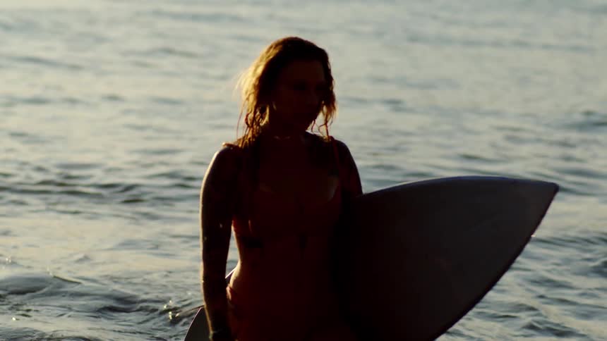 A woman carrying a surfboard walks through shallow water at the beach