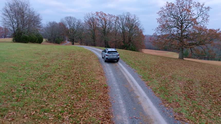 An suv car driving on an asphalt road in a rural area with green fields on the sides and trees with fallen leaves