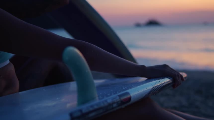 A closeup shot shows a person sitting on a sandy beach holding a surfboard with their hand resting on the boards tail the background features the blurred silhouette of the ocean and the pink and orange colors of a sunset or sunrise