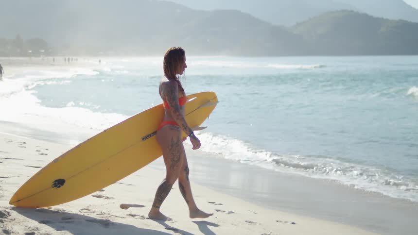 Tattooed woman carries a yellow surfboard while walking on the sand by the sea