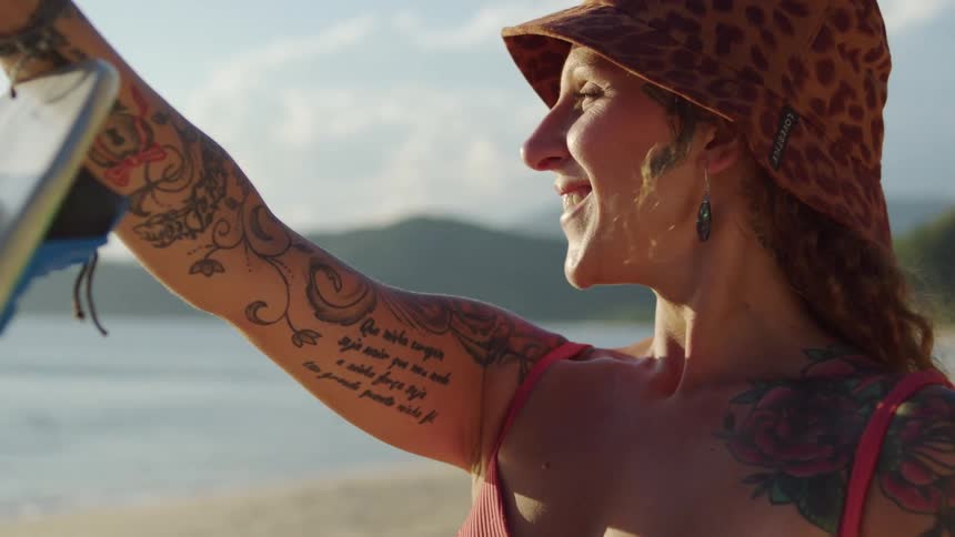 Woman with several tattoos holds a surfboard and smiles while standing on the beach