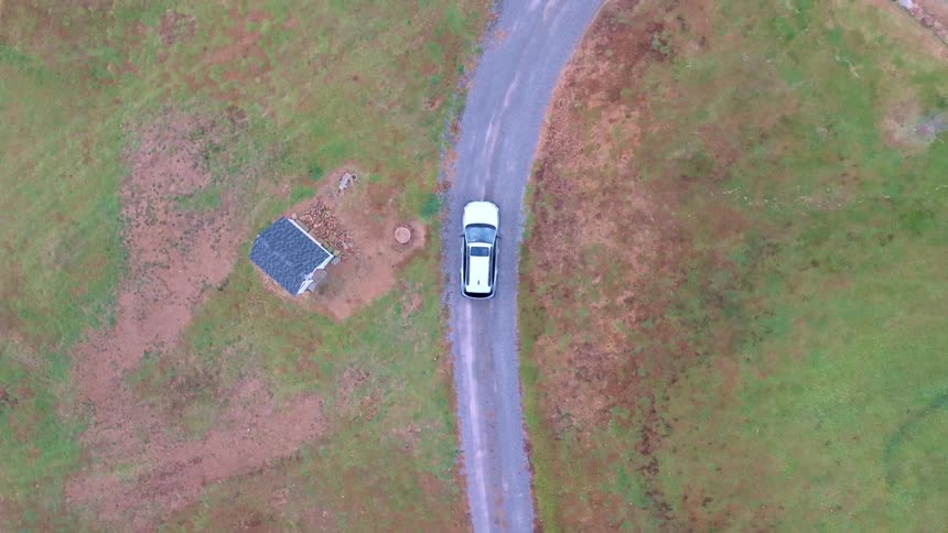 White car seen from above drives along a narrow road surrounded by green and earthy fields