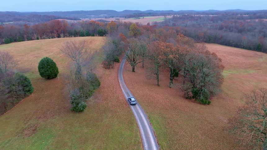 A car drives a winding country road through fields and autumn trees