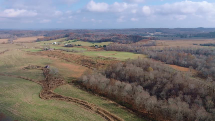An aerial shot captures a rural landscape with rolling hills and patches of deciduous trees in autumn colors a small cluster of buildings is visible in a clearing with a pond nearby under a dramatic cloudy sky