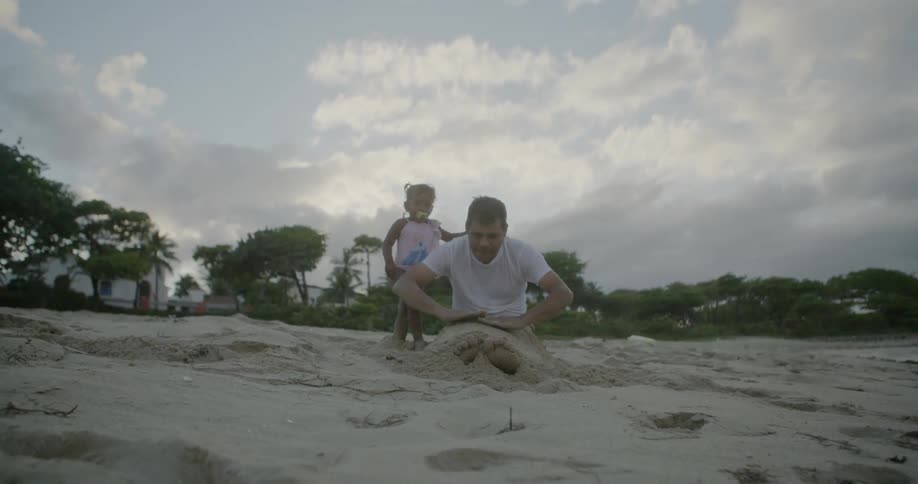 A man buries his legs in beach sand while a toddler watches